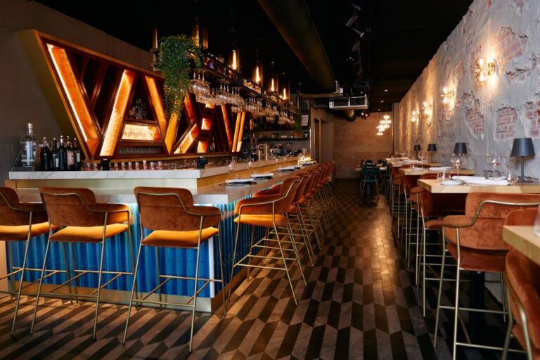Sfeervol modern restaurantinterieur met een lange bar, oranje fluwelen barkrukken en een verlicht houten wanddecor.