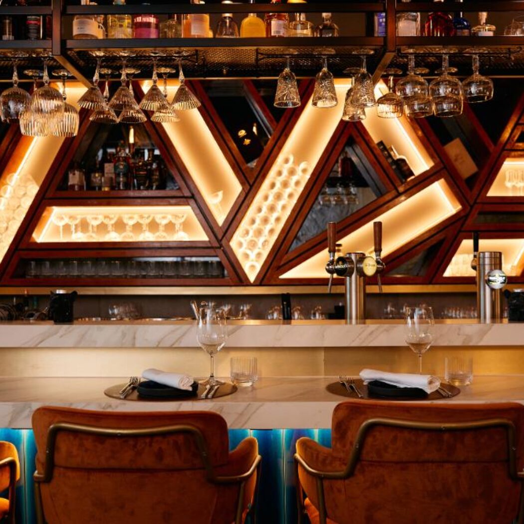 Luxe bar interieur met marmeren toonbank, oranje fluwelen barkrukken en geometrische verlichte drankrekken.