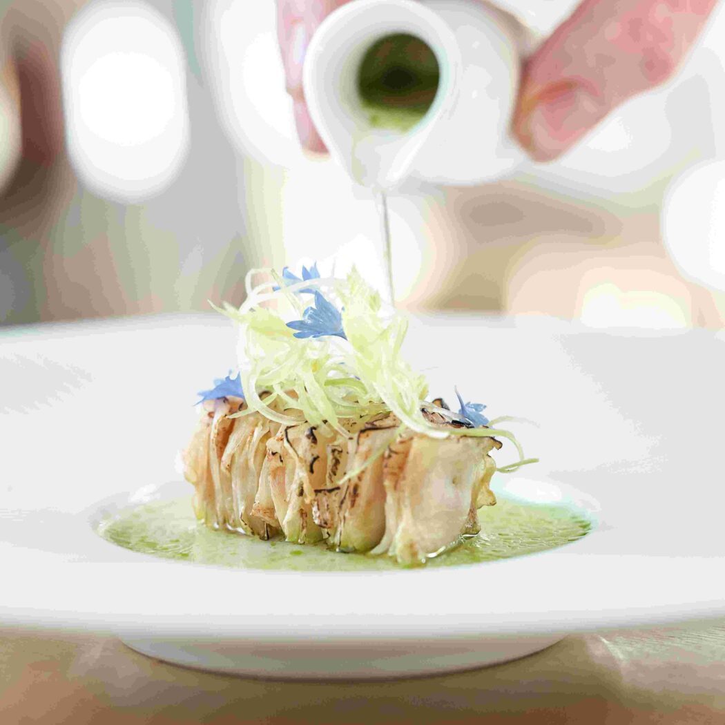 Hand giet groene saus over gegrild gerecht met gevouwen groenten, fijne reepjes en blauwe bloemen.