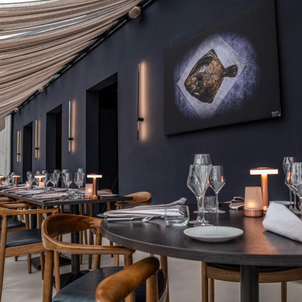 Stijlvol restaurantinterieur met donkere muren, gedrapeerd plafond en gedekte tafels met viskunst.