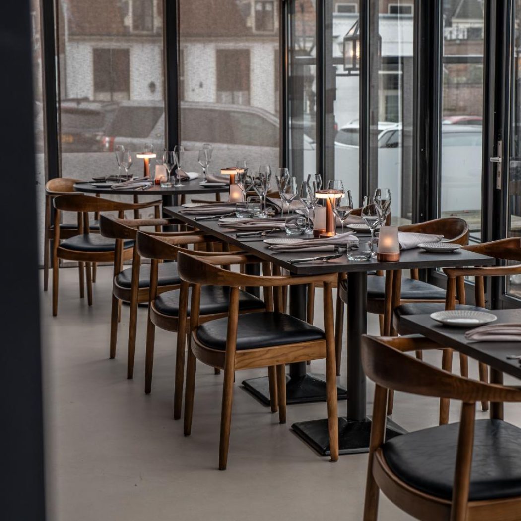 Moderne restaurantruimte met rijen gedekte zwarte tafels, houten stoelen en grote ramen.