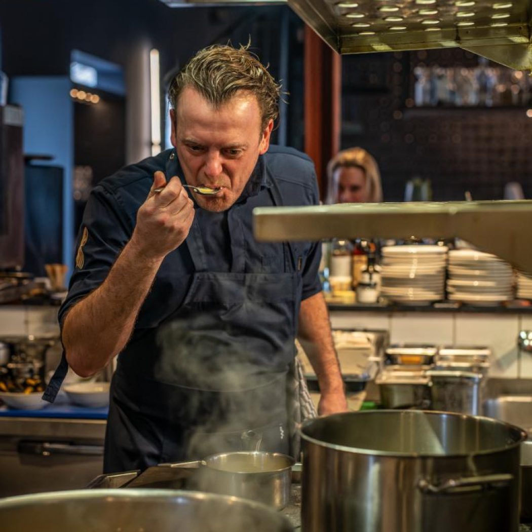 Chefkok proeft aandachtig uit een lepel boven een stomende pan in een professionele keuken.