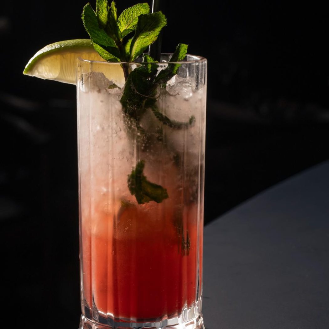 Roze cocktail in een geribbeld glas met munt, limoen en een rietje op een donkere ondergrond.