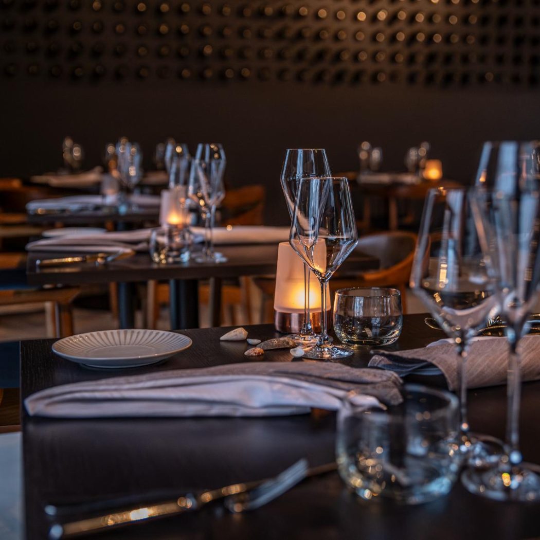 Sfeervolle restauranttafel met wijnglazen, bestek, schelpen en kaarslicht in een elegant donker interieur.