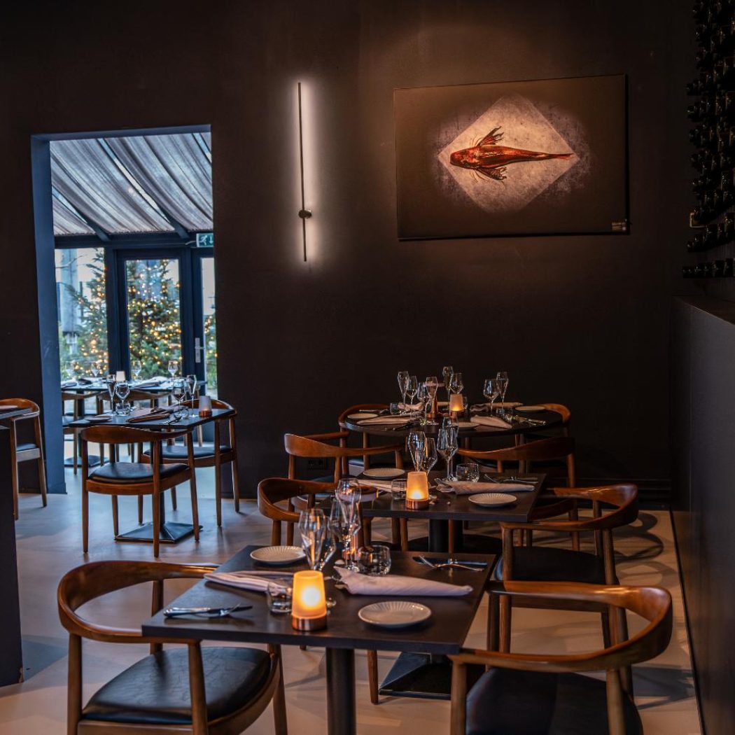 Modern restaurant met donkere muren, gedekte houten tafels, kunstwerk van vis en een muur vol wijnflessen.