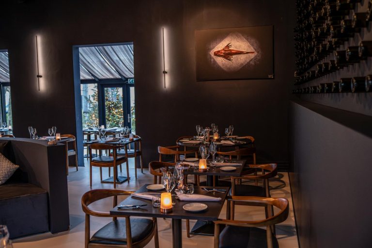Modern restaurant met donkere muren, gedekte houten tafels, kunstwerk van vis en een muur vol wijnflessen.