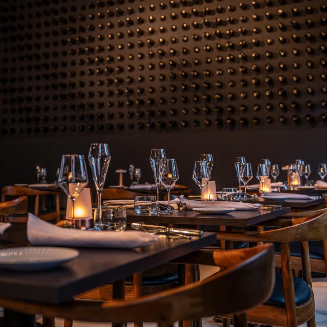 Sfeervol restaurantinterieur met gedekte tafels, wijnglazen en kaarslicht, tegen een donkere muur met wijnflessen.