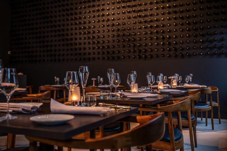 Sfeervol restaurantinterieur met gedekte tafels, wijnglazen en kaarslicht, tegen een donkere muur met wijnflessen.