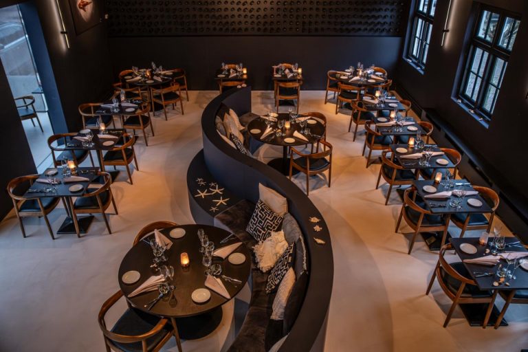 Modern restaurantinterieur met donkere muren, gedekte tafels, houten stoelen en een golvende zitbank.