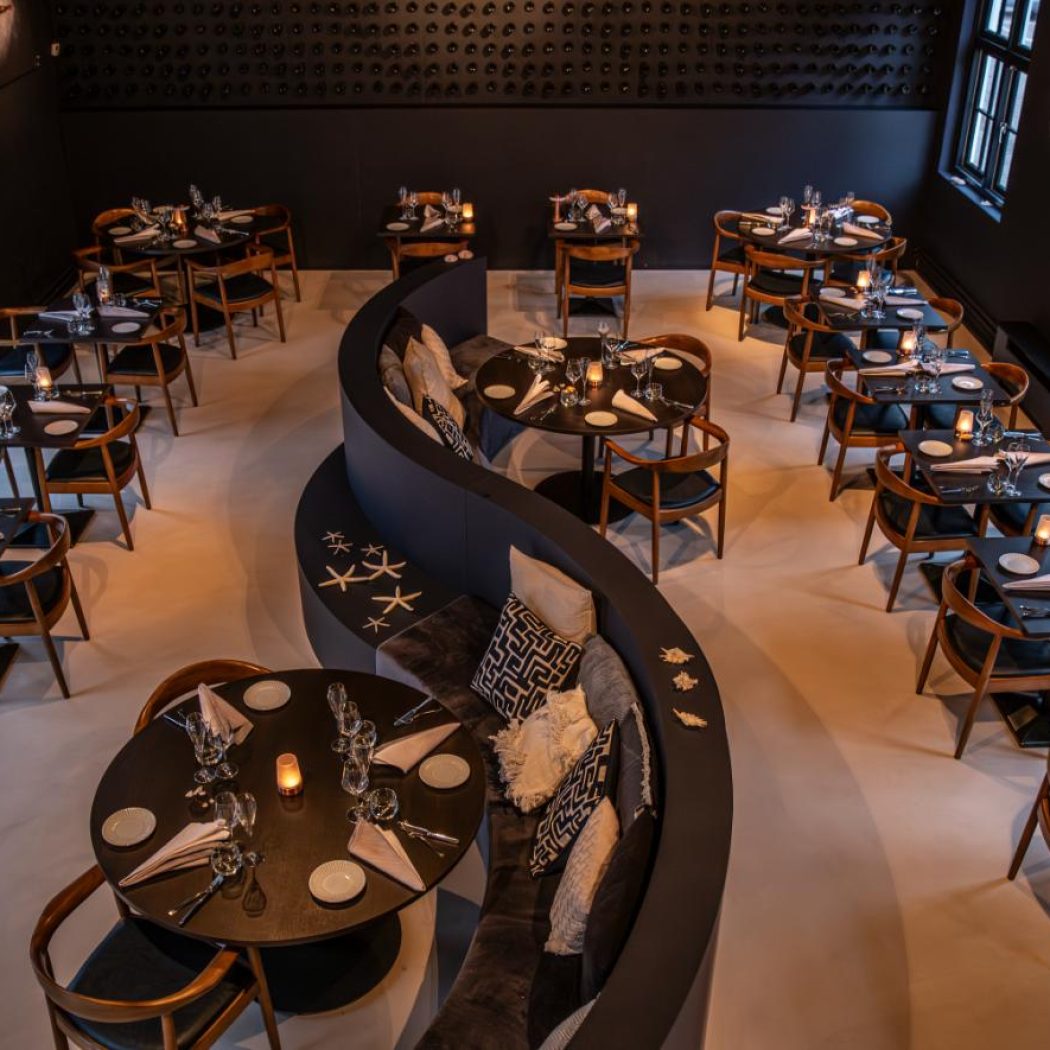 Sfeervol restaurantinterieur met donkere tafels, houten stoelen en een golvende bank met zeesterren.