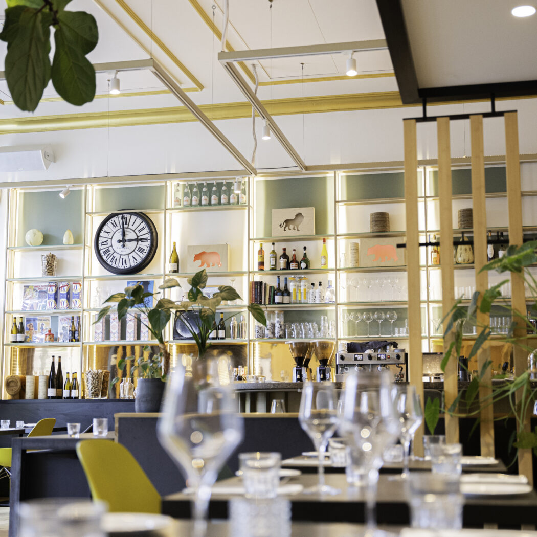 Licht restaurantinterieur met gedekte tafels, een verlichte barwand vol flessen, glazen en een grote klok.