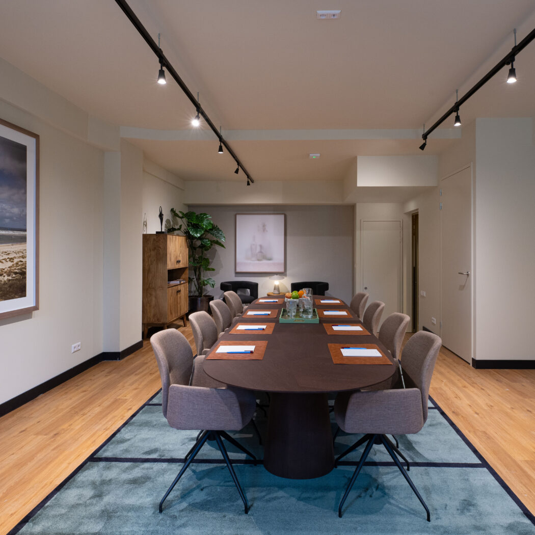 Moderne vergaderzaal met ovale bruine tafel, beige stoelen, turquoise kleed en grote strandfoto.