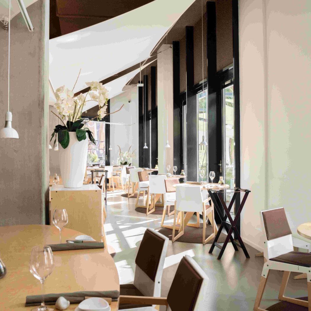 Licht, modern restaurant met houten tafels, witte stoelen, orchideeën en grote ramen.