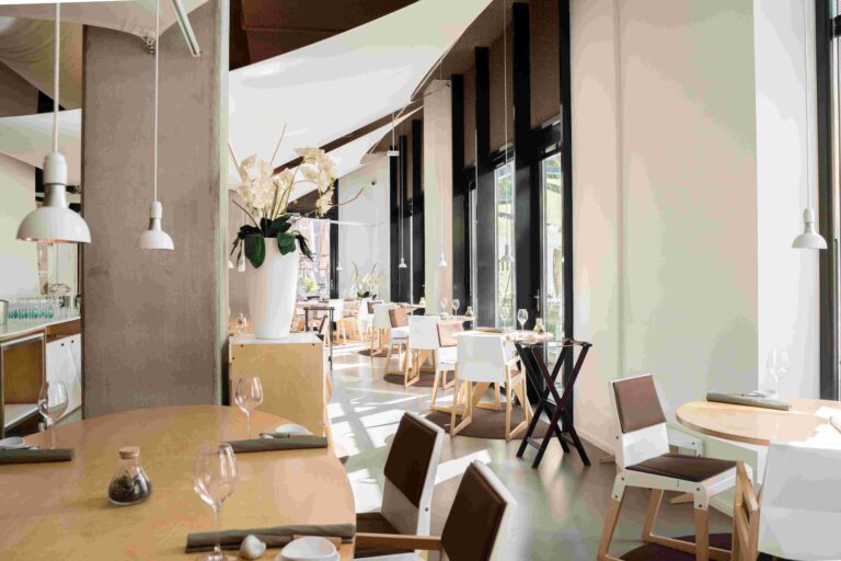 Licht, modern restaurant met houten tafels, witte stoelen, orchideeën en grote ramen.