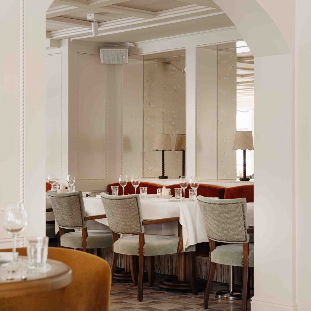 Restaurantinterieur door een lichte boog, met gedekte tafels, grijs gestoffeerde stoelen en rode banken langs spiegelwanden met lampen.