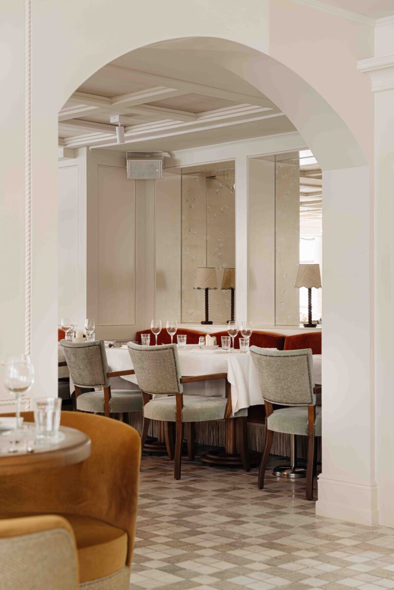 Restaurantinterieur door een lichte boog, met gedekte tafels, grijs gestoffeerde stoelen en rode banken langs spiegelwanden met lampen.