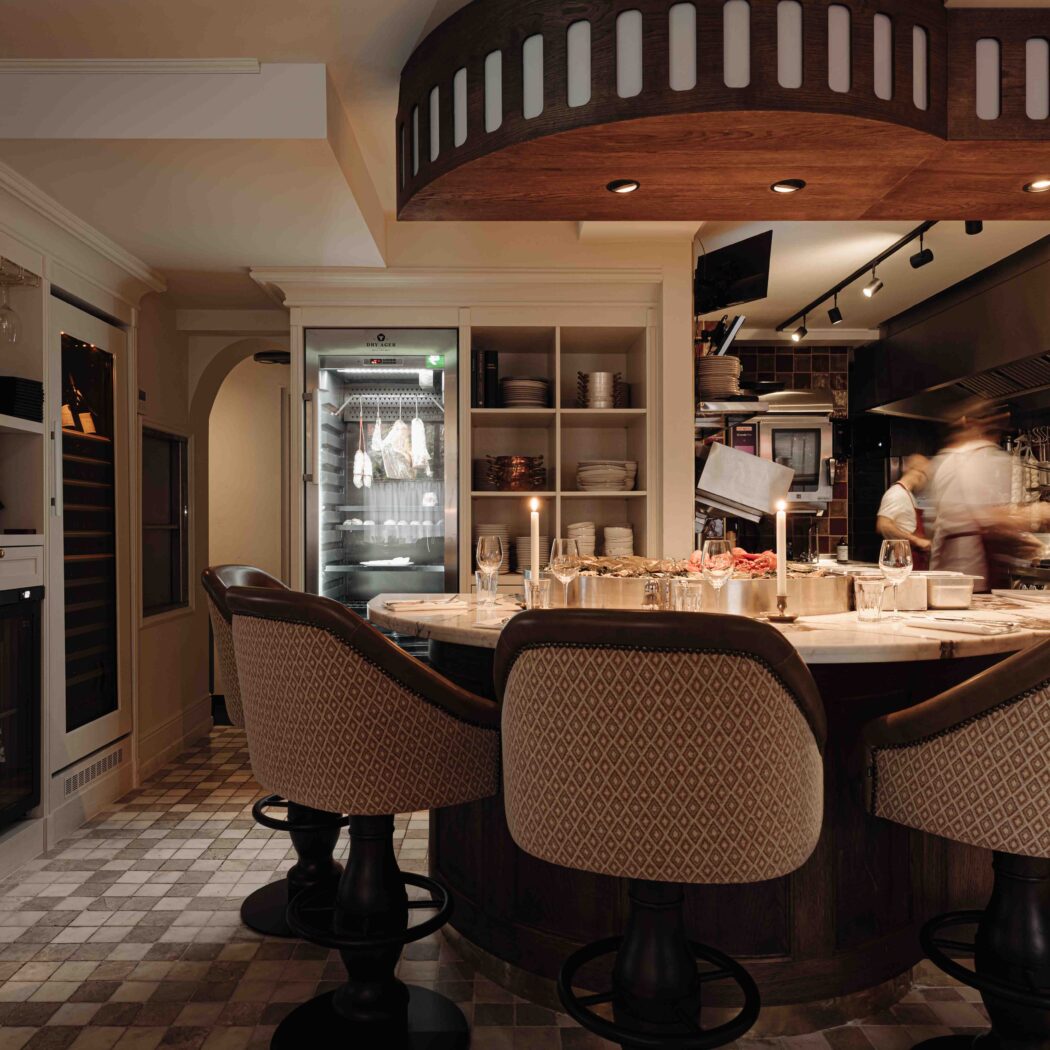 Drie elegante barstoelen aan een marmeren bar in een modern restaurant met open keuken en dry-aging kast.