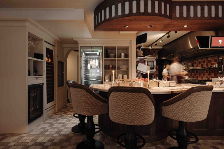 Drie elegante barstoelen aan een marmeren bar in een modern restaurant met open keuken en dry-aging kast.