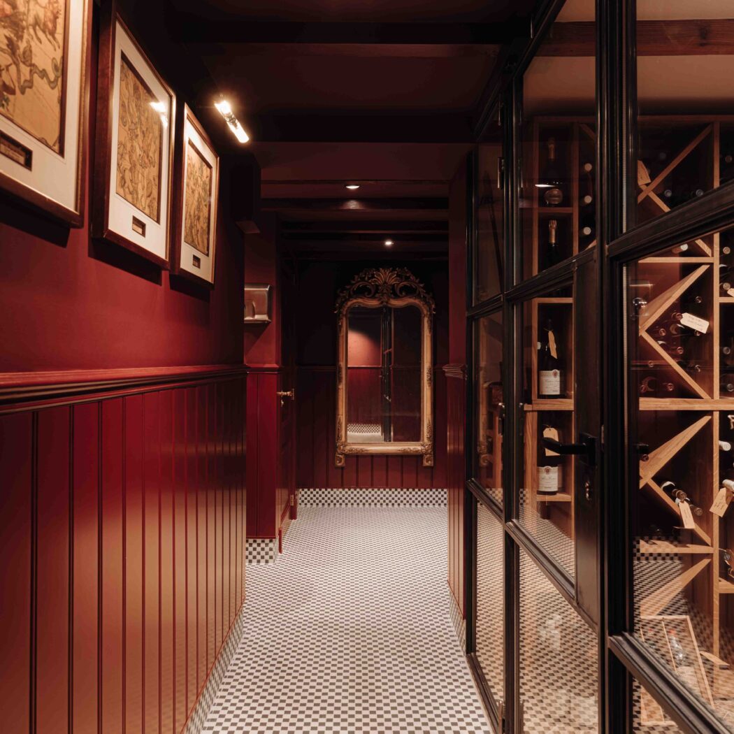 Donkerrode gang met zwart-wit geblokte vloer, sierlijke gouden spiegel en glazen wand naar een wijnkelder.