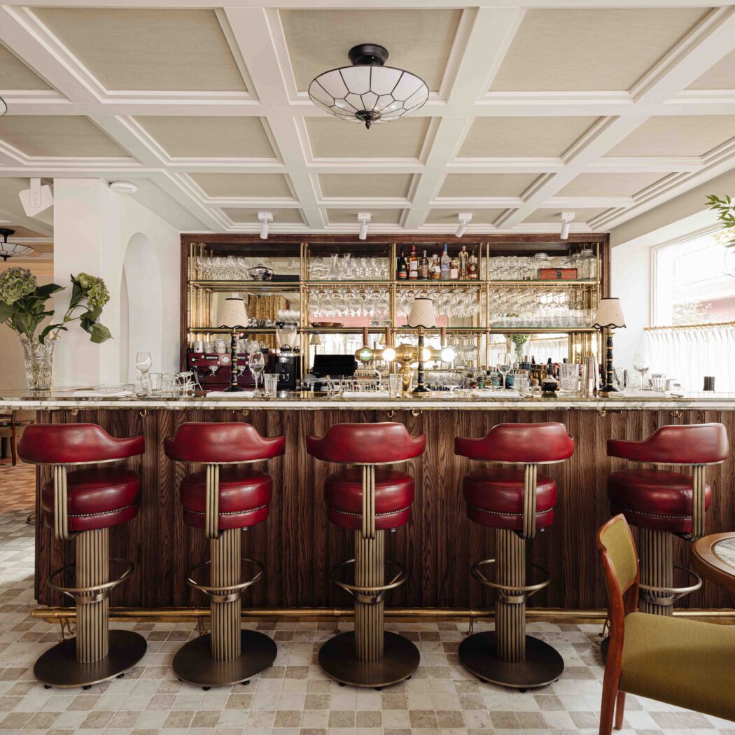 Elegante restaurantbar met marmeren blad, vijf rode leren barkrukken en een gouden achterwand vol flessen en glazen.