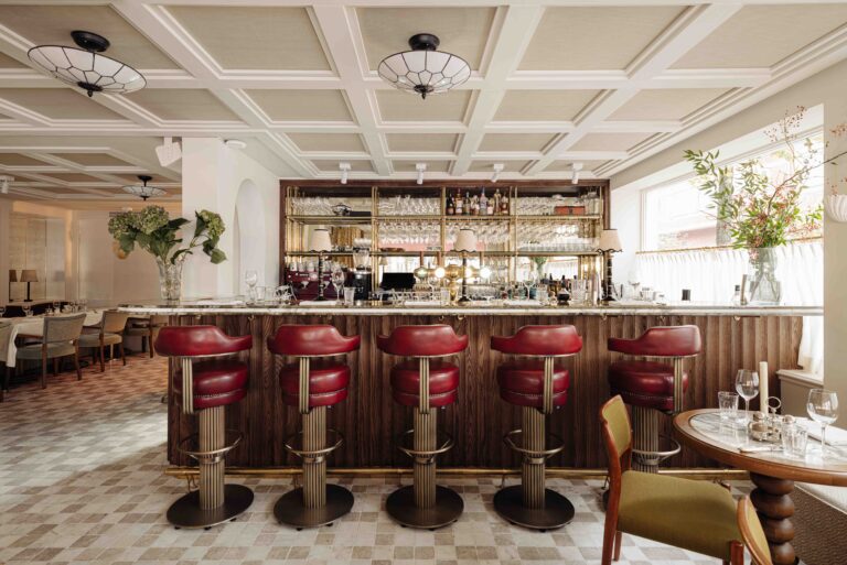 Elegante restaurantbar met marmeren blad, vijf rode leren barkrukken en een gouden achterwand vol flessen en glazen.