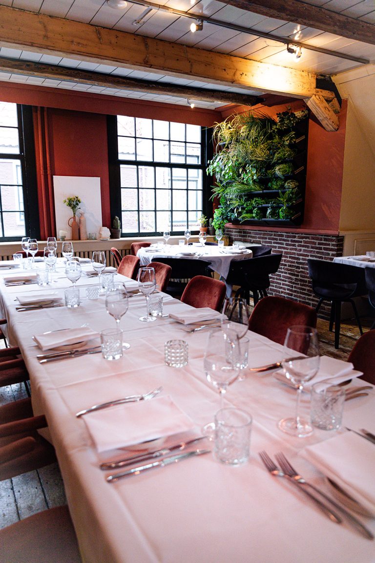 Gedekte eettafel in een sfeervol restaurant met velvet stoelen, houten balken en een groene plantenwand.