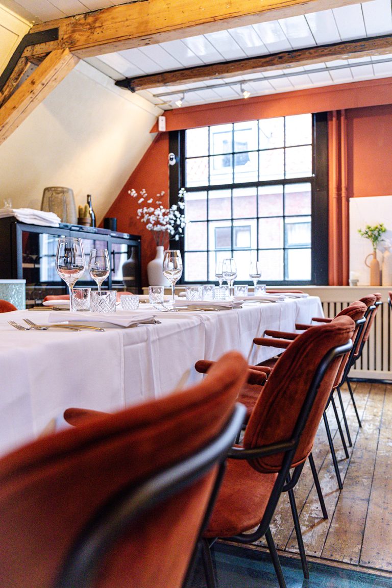 Lange gedekte tafel met witte linnen en glazen, geflankeerd door terracotta stoelen, in een restaurantruimte met houten balken.