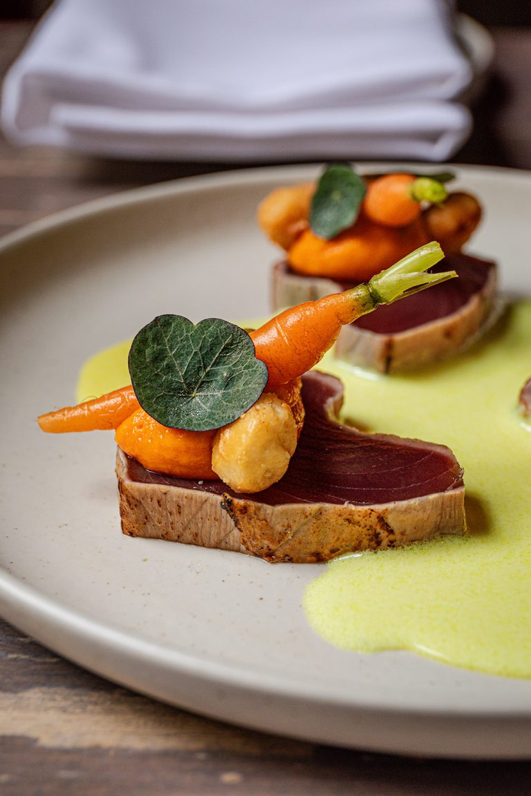 Culinair gerecht van rosé gebakken tonijn met babywortels, verse garnituur en een limoengroene saus op een beige bord.