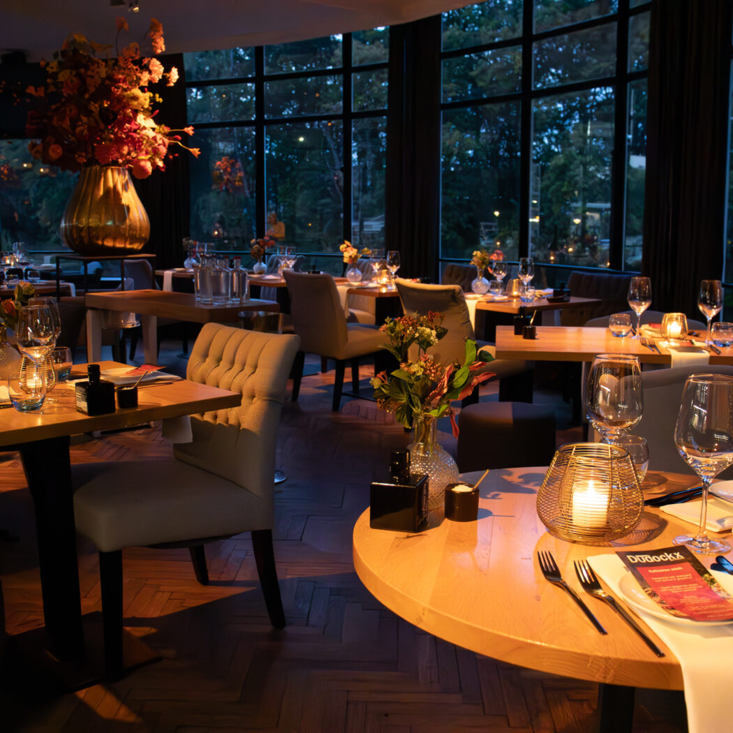 Sfeervol restaurant interieur met gedekte houten tafels, kaarsen en DBBoekX menus, uitzicht op bos bij schemering.