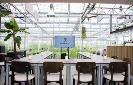 Moderne evenementenlocatie in glazen kas met lange houten tafels, stoelen, bloemen en een scherm met STADSJOCHIES.