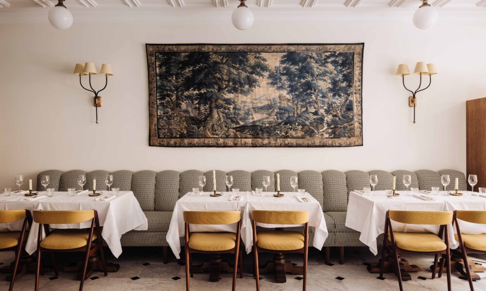 Stylish restaurant interior with a large landscape tapestry, a striped sofa and tables with yellow chairs.