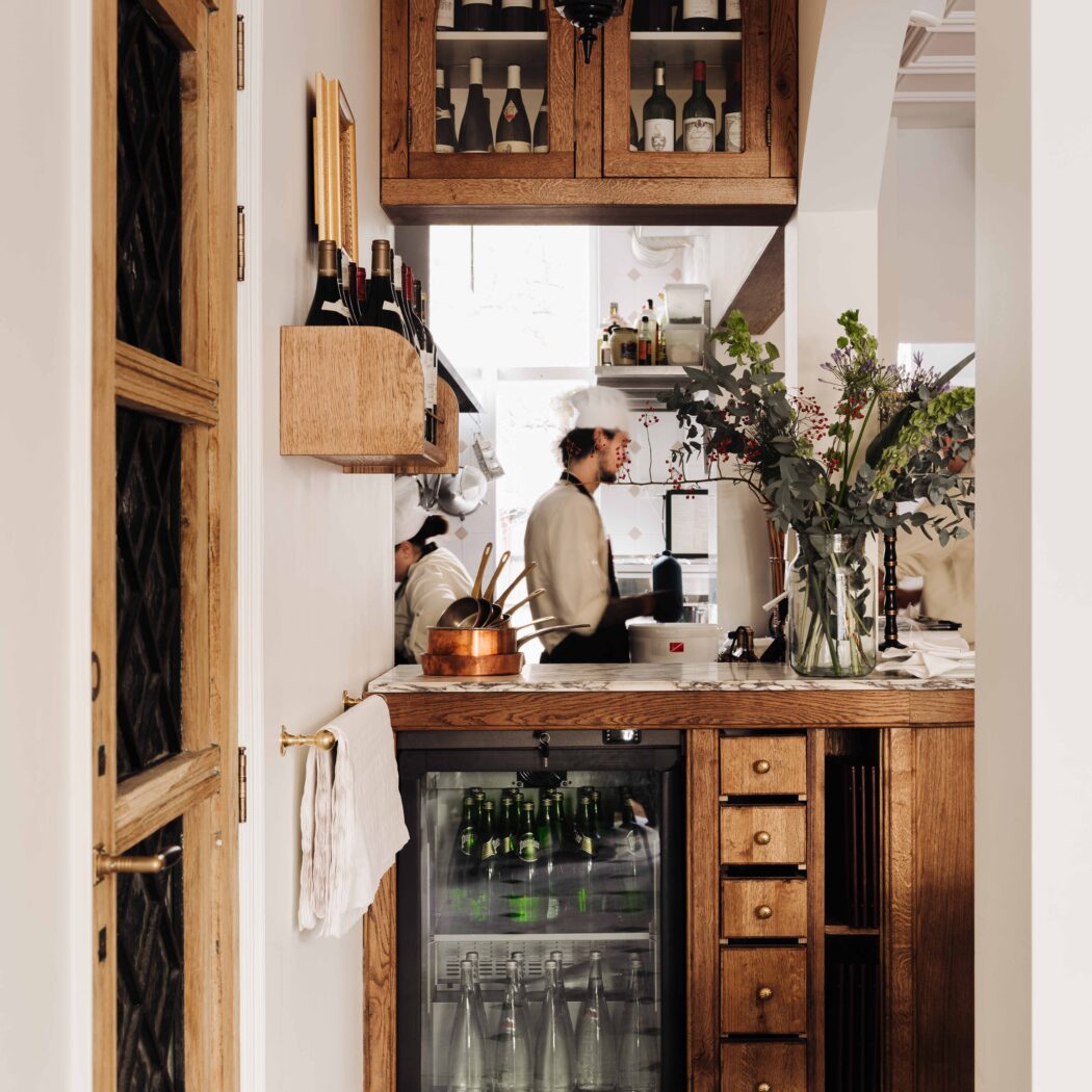 Klassieke restaurantbar met houten kasten vol wijn, een koelkast met drank, en chefs aan het werk in de open keuken.