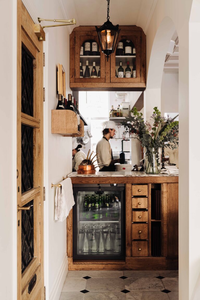 Klassieke restaurantbar met houten kasten vol wijn, een koelkast met drank, en chefs aan het werk in de open keuken.