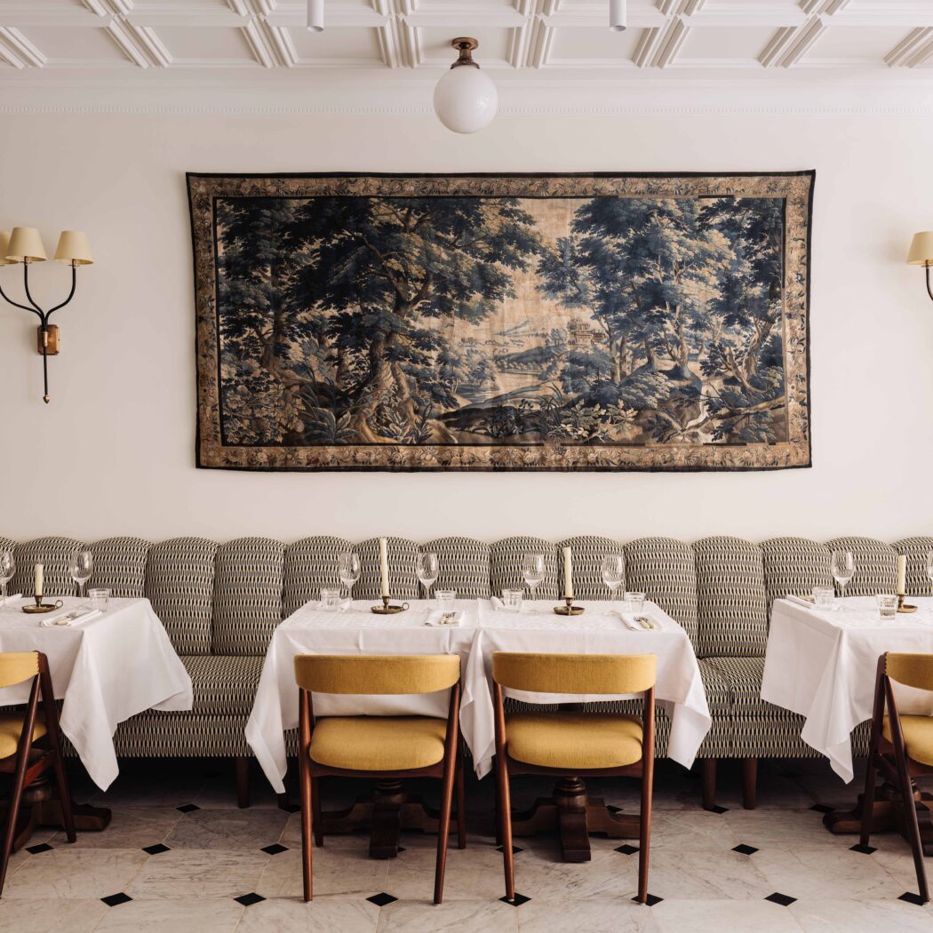 Stijlvol restaurantinterieur met een groot landschaps-wandtapijt, een gestreepte bank en tafels met gele stoelen.