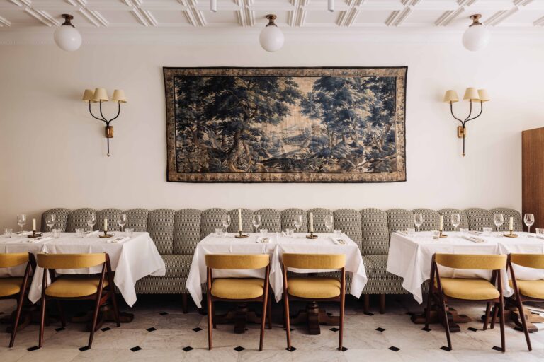 Stijlvol restaurantinterieur met een groot landschaps-wandtapijt, een gestreepte bank en tafels met gele stoelen.