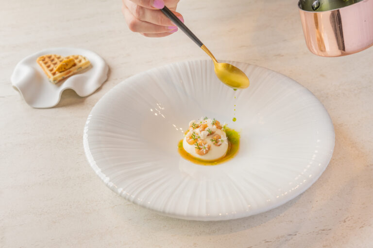 Goudkleurige lepel schenkt groene saus over een wit culinair gerecht met garnalen en kruiden, geserveerd op een wit bord.