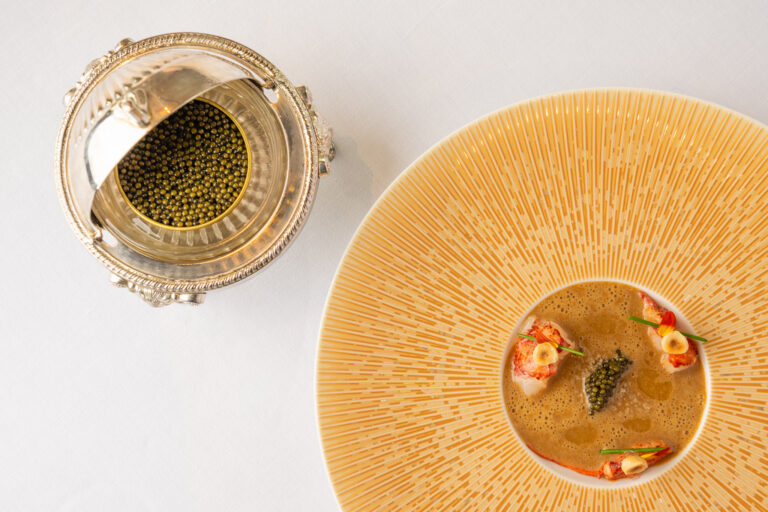 Decoratieve zilveren kaviaarhouder met kaviaar en een gouden bord met kreeft, kaviaar en bieslook in bruine bouillon.