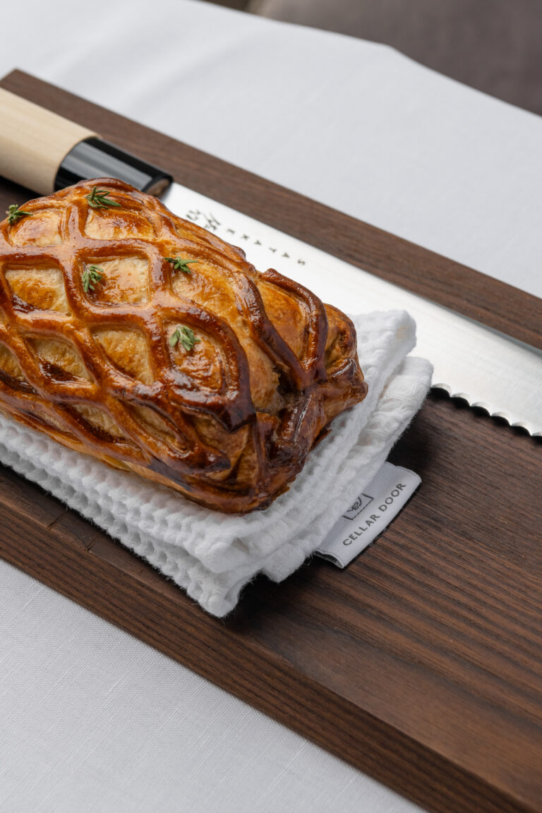 Goudbruin roosterbladerdeeg gebak met verse kruiden op een witte doek met ‘CELLAR DOOR’, naast een broodmes op een houten plank.