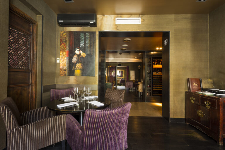 Restaurant met gedekte ronde tafel, paarse fluwelen stoelen, Aziatische kunst en doorkijk naar wijnrek.