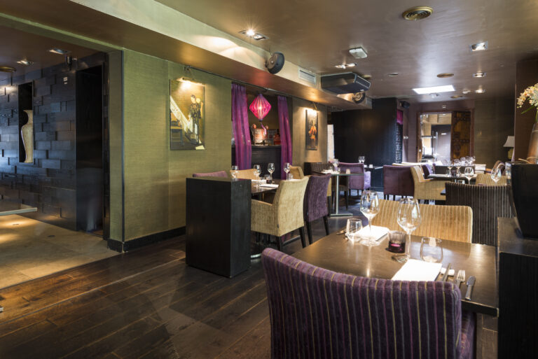 Sfeervol restaurant interieur met donkere houten vloer, groene muren, paarse en beige stoelen en gedekte tafels.