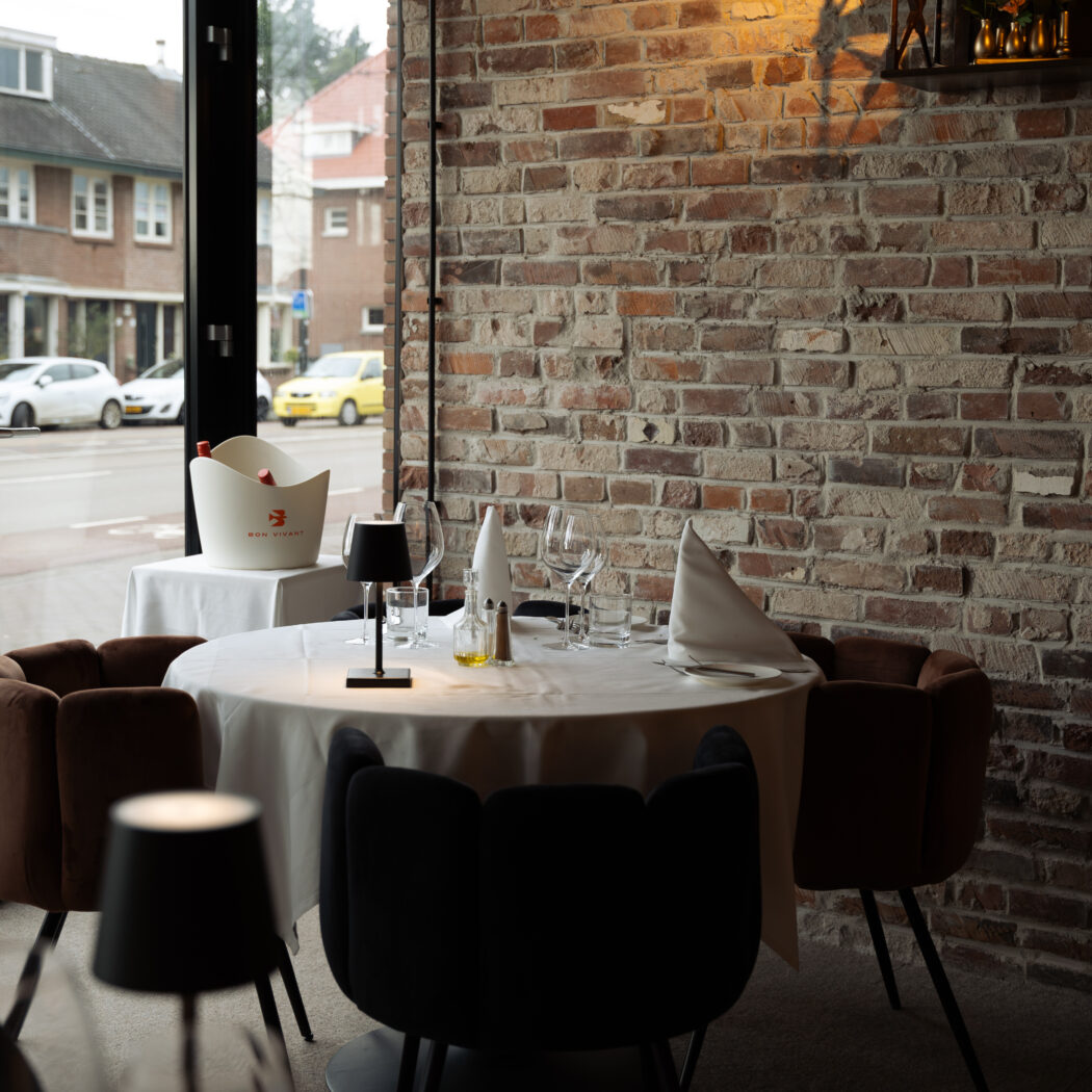 Gedekte restauranttafel met wijnglazen, een bakstenen muur en een BON VIVANT wijnkoeler bij het raam.