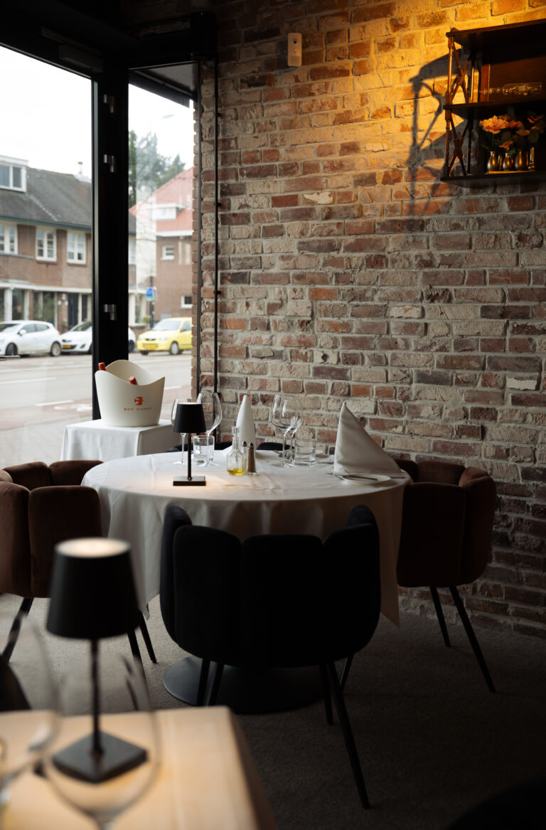 Gedekte restauranttafel met wijnglazen, een bakstenen muur en een BON VIVANT wijnkoeler bij het raam.