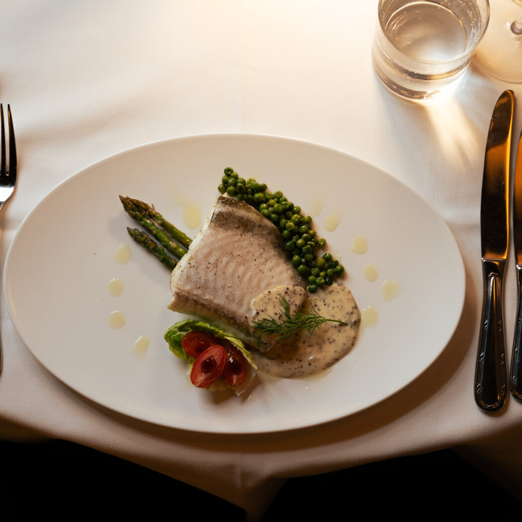 Visfilet met groene peperkorrels, asperges, dillesaus en tomatensalade op wit bord met bestek en glazen.