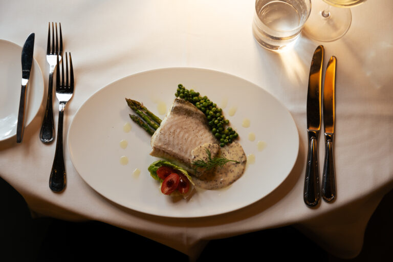 Visfilet met groene peperkorrels, asperges, dillesaus en tomatensalade op wit bord met bestek en glazen.