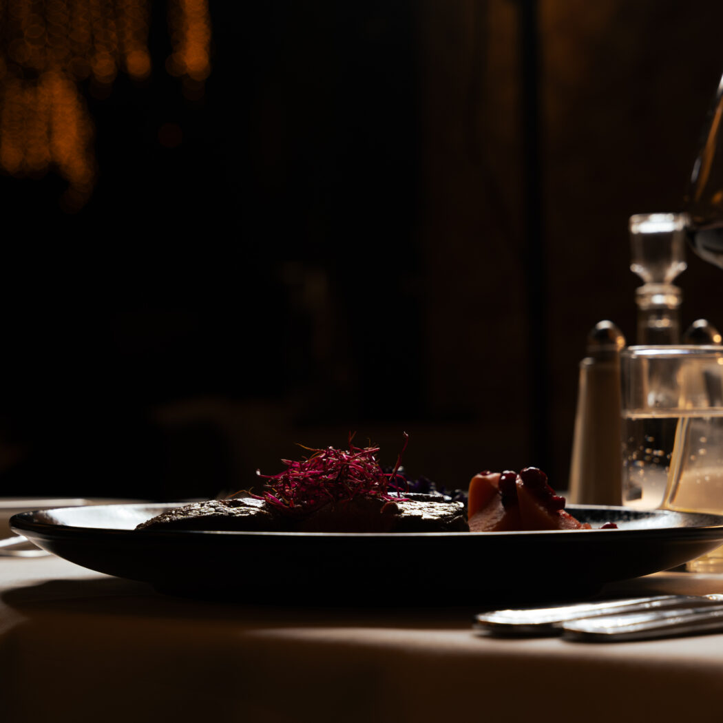 Een elegant gerecht met rode kiemen op een donker bord, naast een wijnglas en water in een sfeervolle setting.