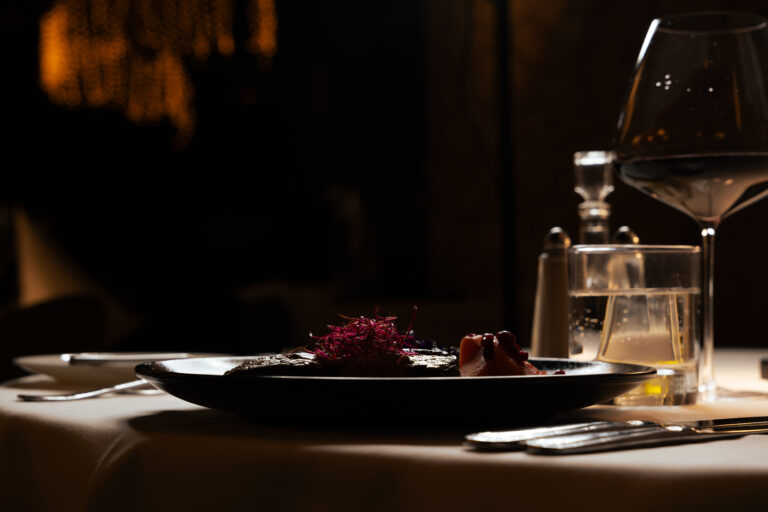 Een elegant gerecht met rode kiemen op een donker bord, naast een wijnglas en water in een sfeervolle setting.
