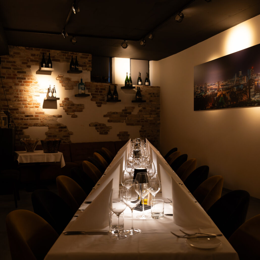 Intieme dinerruimte met lange, feestelijk gedekte tafel, wijnglazen, bakstenen muur en verlicht stadsgezicht.