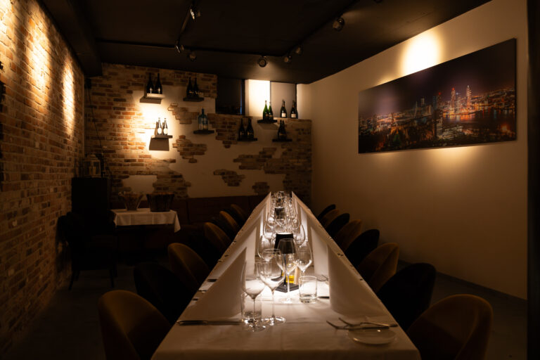 Intieme dinerruimte met lange, feestelijk gedekte tafel, wijnglazen, bakstenen muur en verlicht stadsgezicht.