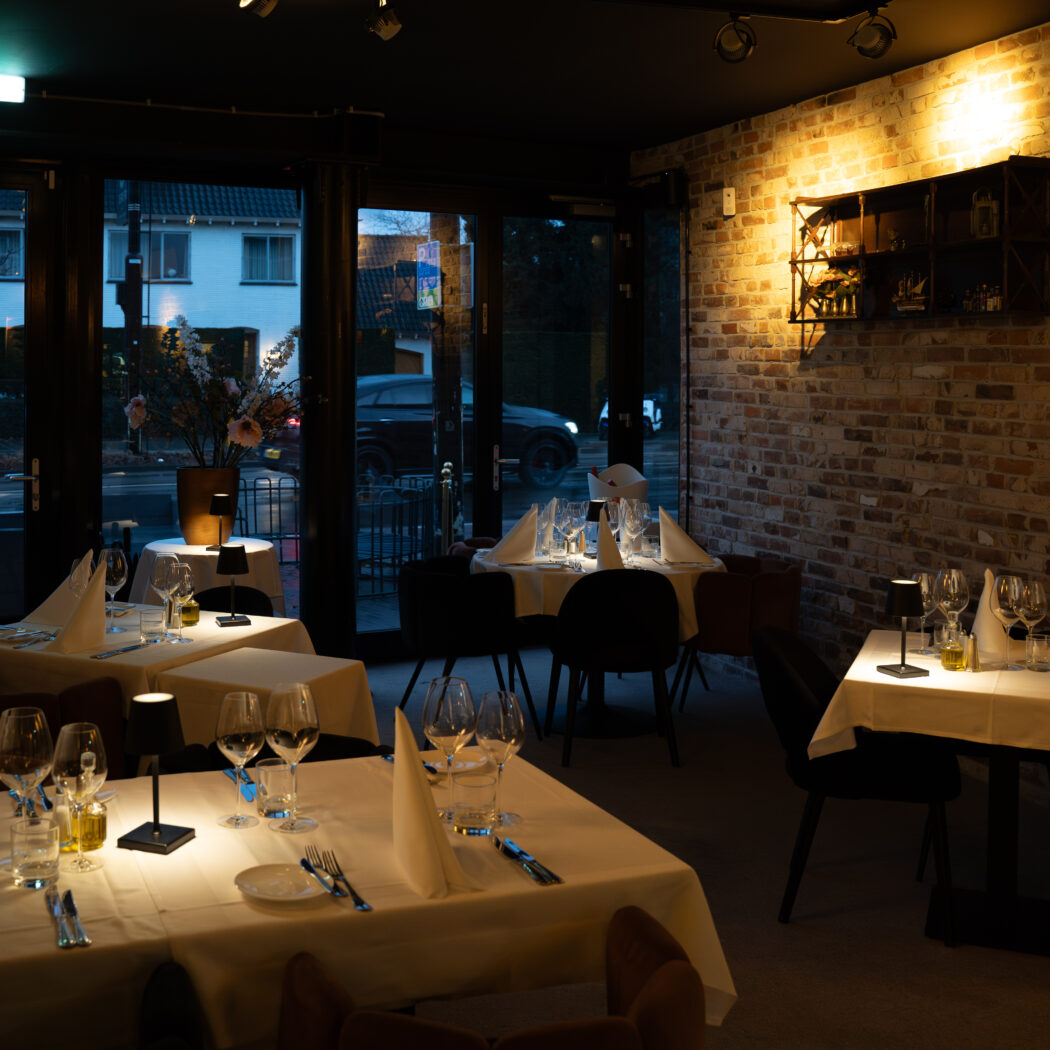 Warm verlicht restaurantinterieur met meerdere gedekte tafels, kleine lampjes, een bakstenen muur en uitzicht op de straat.