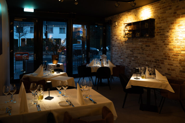 Warm verlicht restaurantinterieur met meerdere gedekte tafels, kleine lampjes, een bakstenen muur en uitzicht op de straat.
